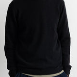 A man wearing a black crewneck knit sweater, paired with dark blue corduroy trousers and a light beige undershirt, shown from the chin down against a plain white wall.