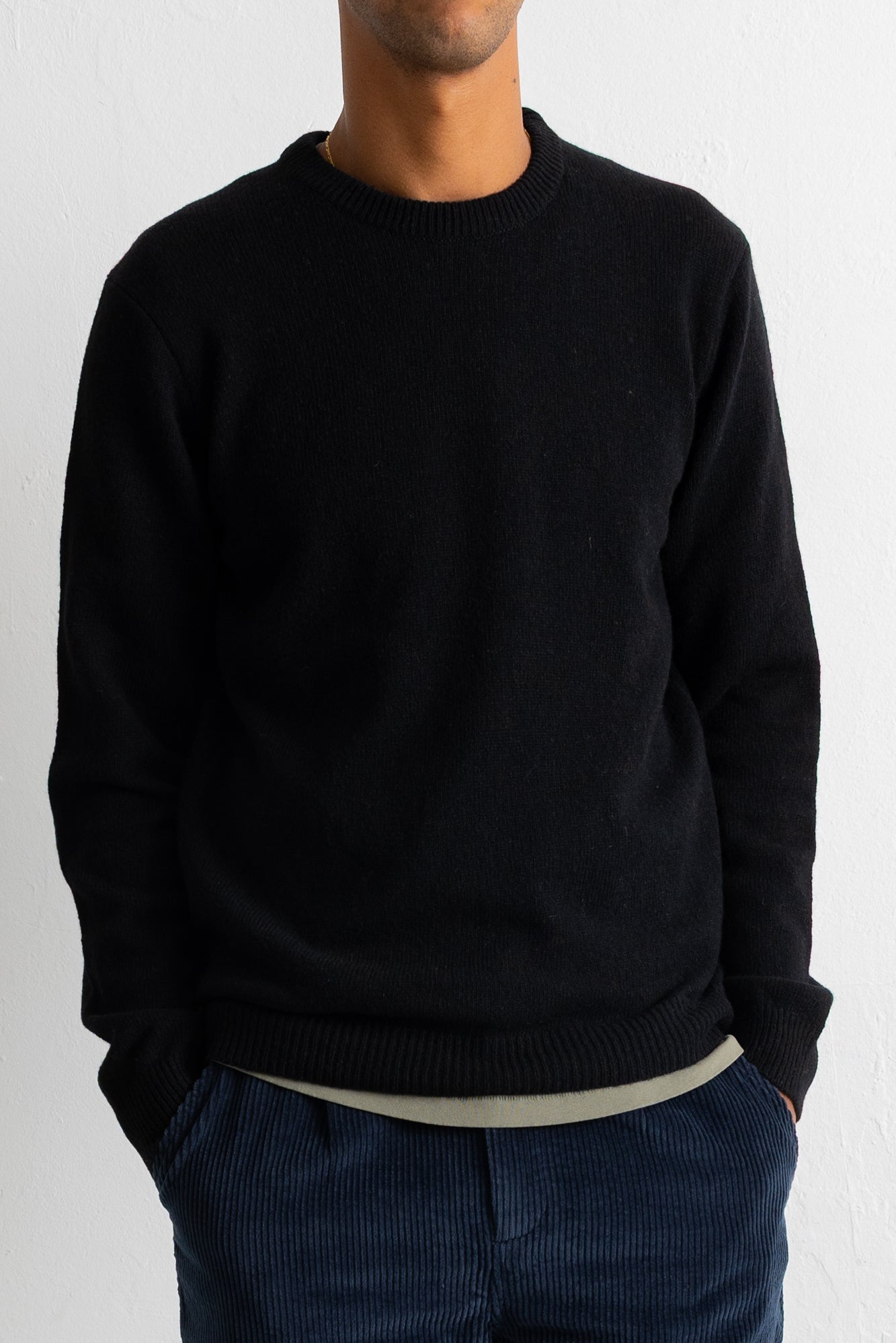 A man wearing a black crewneck knit sweater, paired with dark blue corduroy trousers and a light beige undershirt, shown from the chin down against a plain white wall.