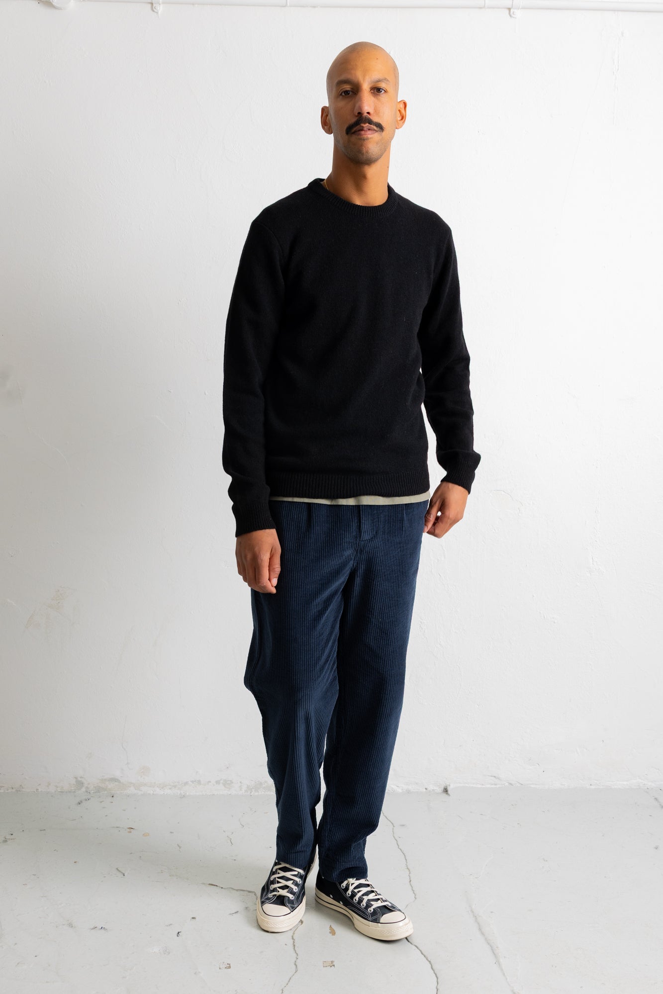 A man wearing a black crewneck sweatshirt, dark blue baggy pants, and black and white canvas sneakers, standing against a white studio wall.
