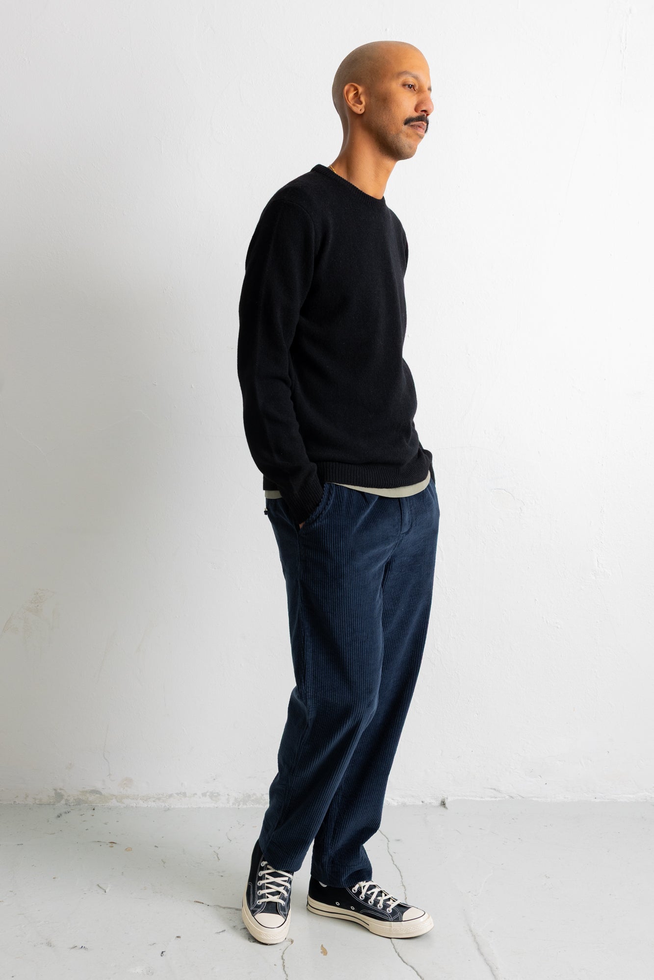 A man standing in profile wearing a black long-sleeve crewneck sweater, dark blue corduroy trousers, and black canvas sneakers against a plain white studio background.