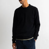 A man wearing a black crewneck sweater and dark blue corduroy trousers, standing against a plain white wall.