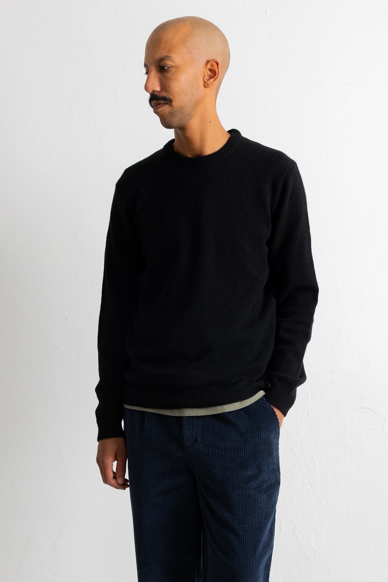 A man wearing a black crewneck sweater and dark blue corduroy trousers, standing against a plain white wall.