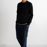A man wearing a black crewneck sweater, dark blue jeans, and navy sneakers with white soles, standing against a white studio wall.