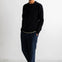 A man wearing a black crewneck sweater, dark blue jeans, and navy sneakers with white soles, standing against a white studio wall.