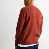 A man wearing a rust-orange knit crewneck sweater, shown from a rear three-quarter angle against a plain white studio background.