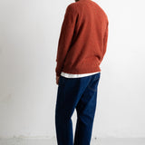 A man wearing a rust-orange knit sweater, dark blue jeans, and black canvas sneakers, shown from a side-back angle against a plain white studio wall.