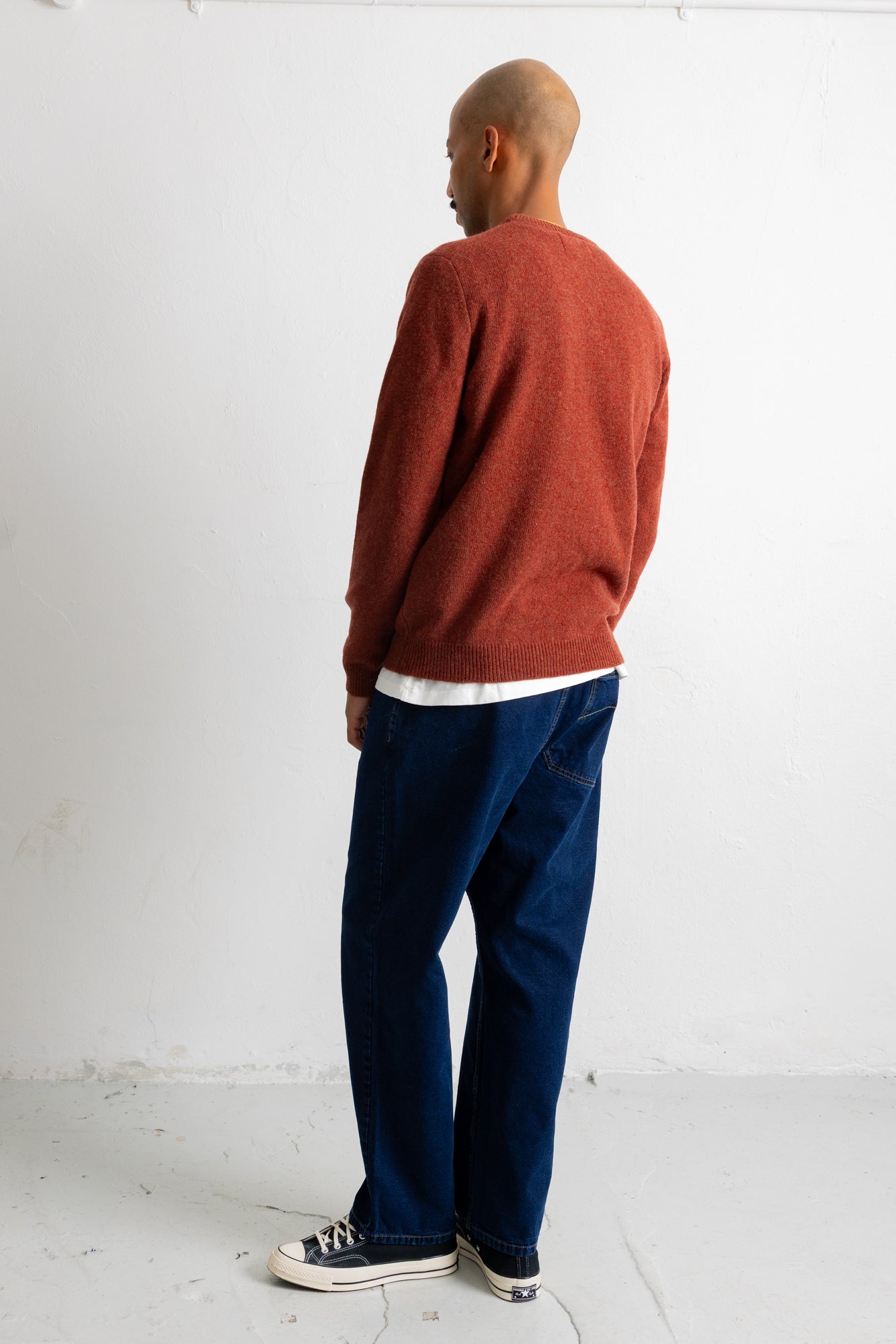 A man wearing a rust-orange knit sweater, dark blue jeans, and black canvas sneakers, shown from a side-back angle against a plain white studio wall.