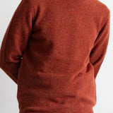 A man wearing a rust-colored crewneck knit sweater, paired with a white undershirt and blue jeans, shown from the neck down against a plain white wall.