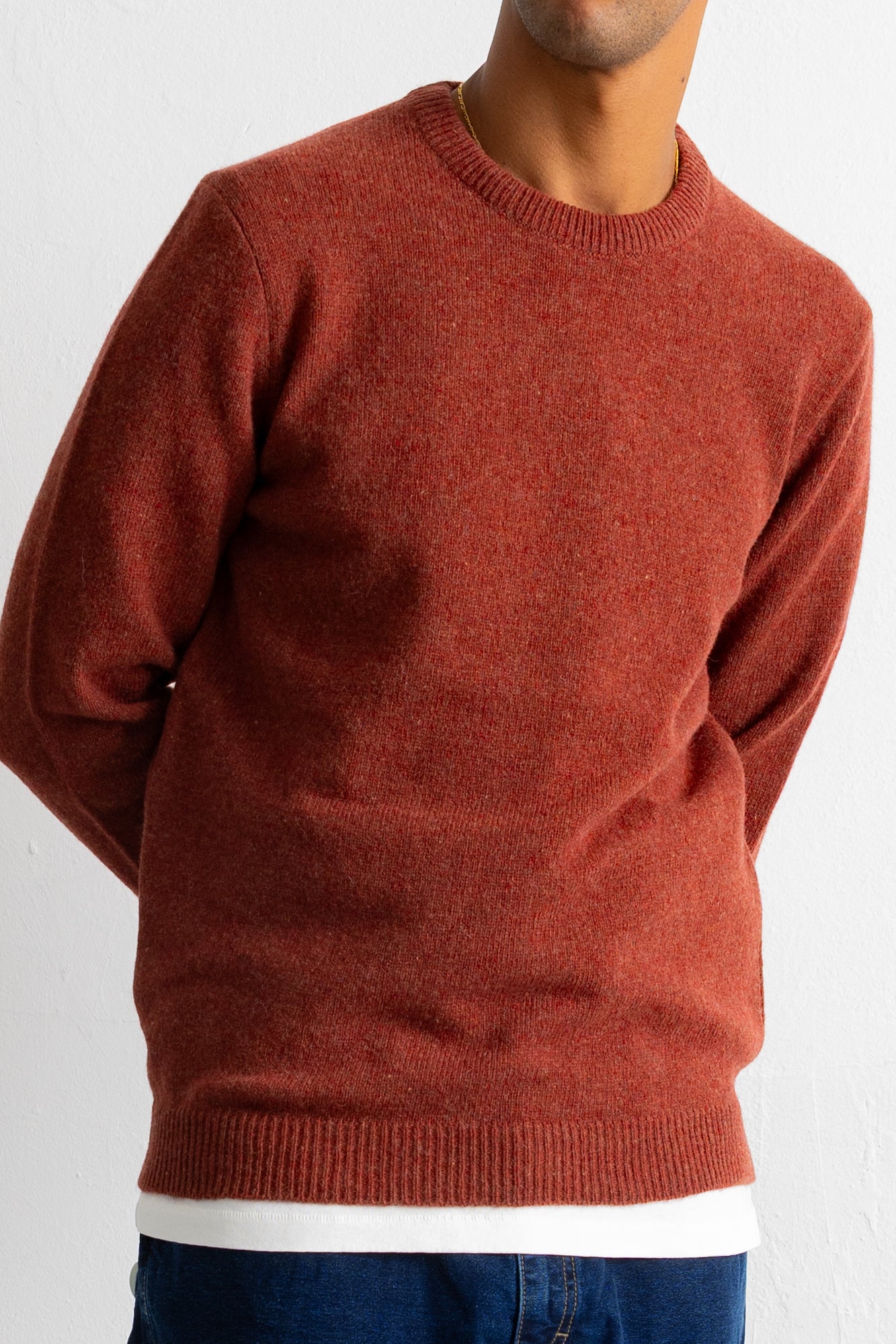 A man wearing a rust-colored crewneck knit sweater, paired with a white undershirt and blue jeans, shown from the neck down against a plain white wall.