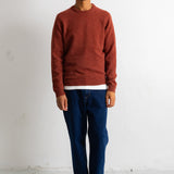 A man wearing a rust-colored crewneck sweater, dark blue jeans, and black-and-white sneakers, standing against a plain white wall.