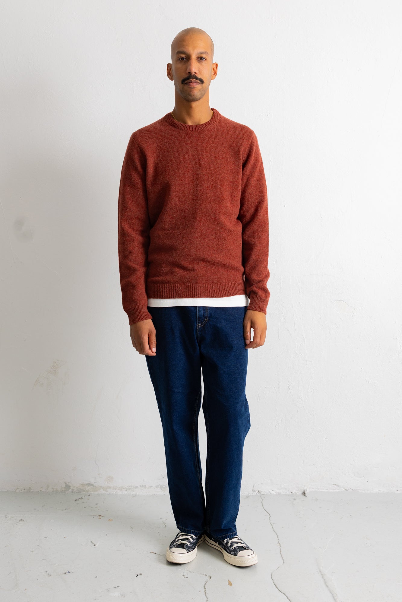 A man wearing a rust-colored crewneck sweater, dark blue jeans, and black-and-white sneakers, standing against a plain white wall.