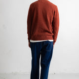 A man viewed from behind wearing a rust-colored knit sweater, dark blue jeans, and black-and-white sneakers, standing against a plain white wall.