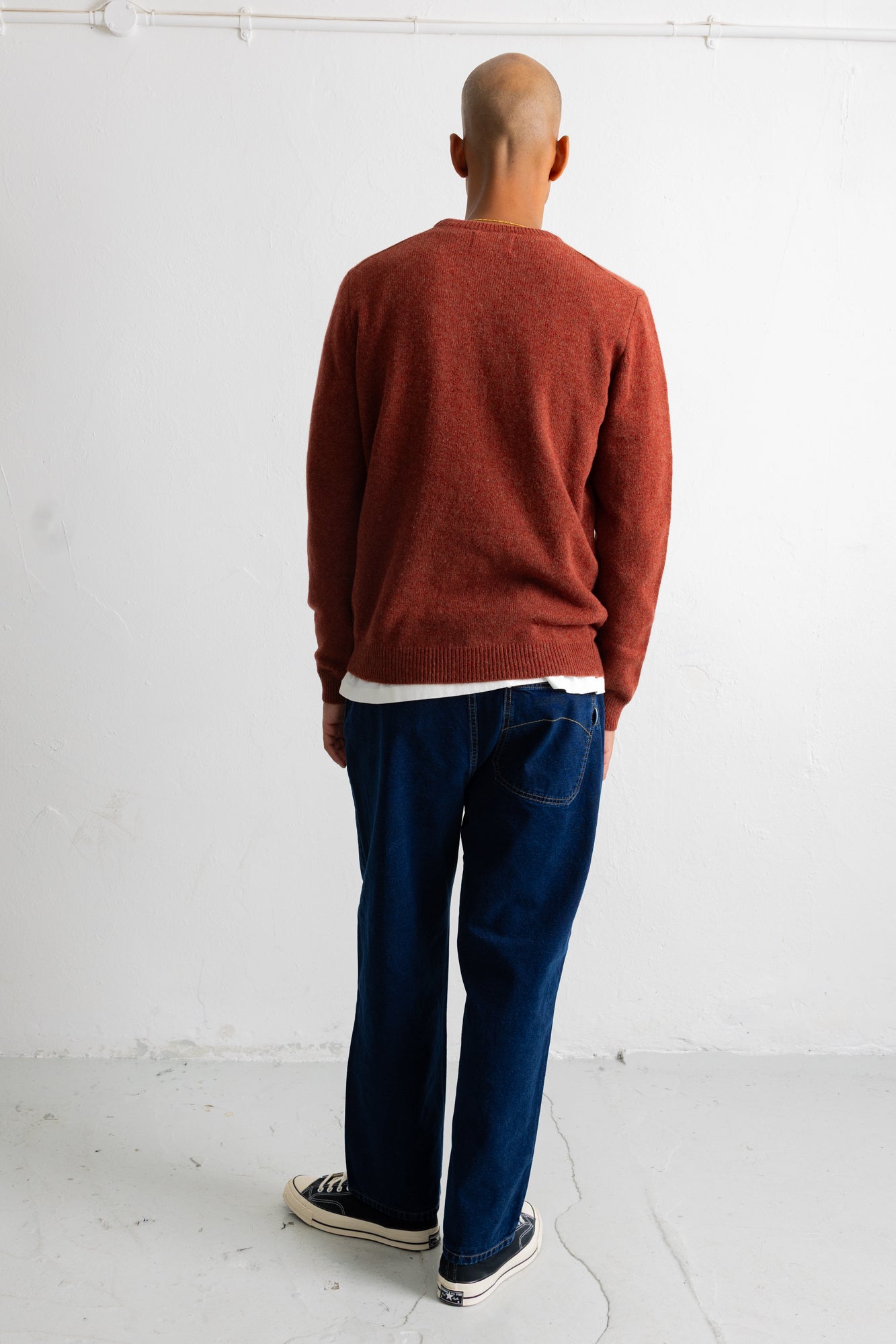 A man viewed from behind wearing a rust-colored knit sweater, dark blue jeans, and black-and-white sneakers, standing against a plain white wall.