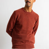 A man wearing a rust-colored crewneck knit sweater over a white undershirt and blue jeans, standing against a plain white wall.