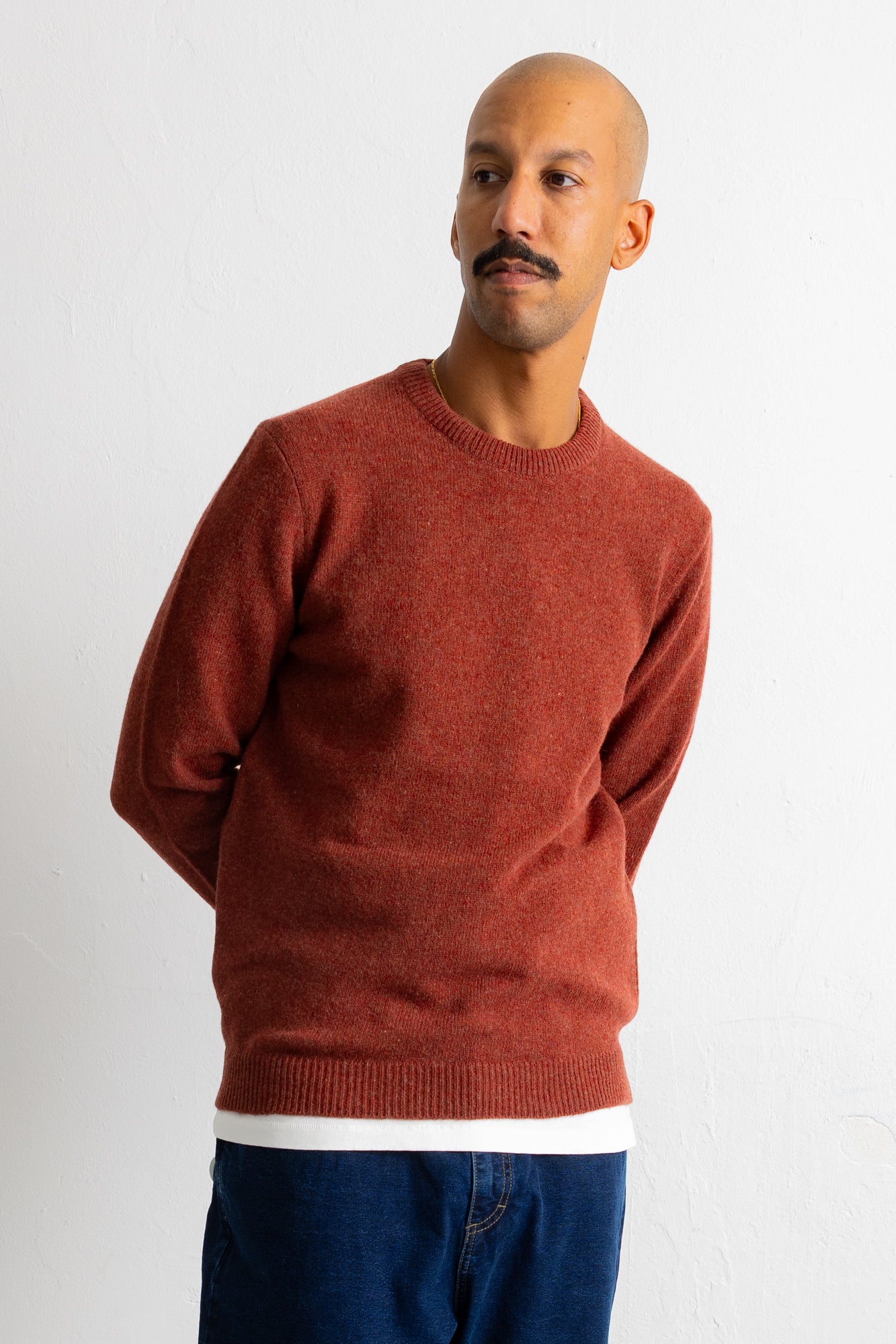 A man wearing a rust-colored crewneck knit sweater over a white undershirt and blue jeans, standing against a plain white wall.