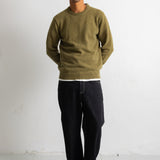 A man wearing an olive green crewneck sweater, black pants, and black-and-white sneakers, standing against a plain white wall.