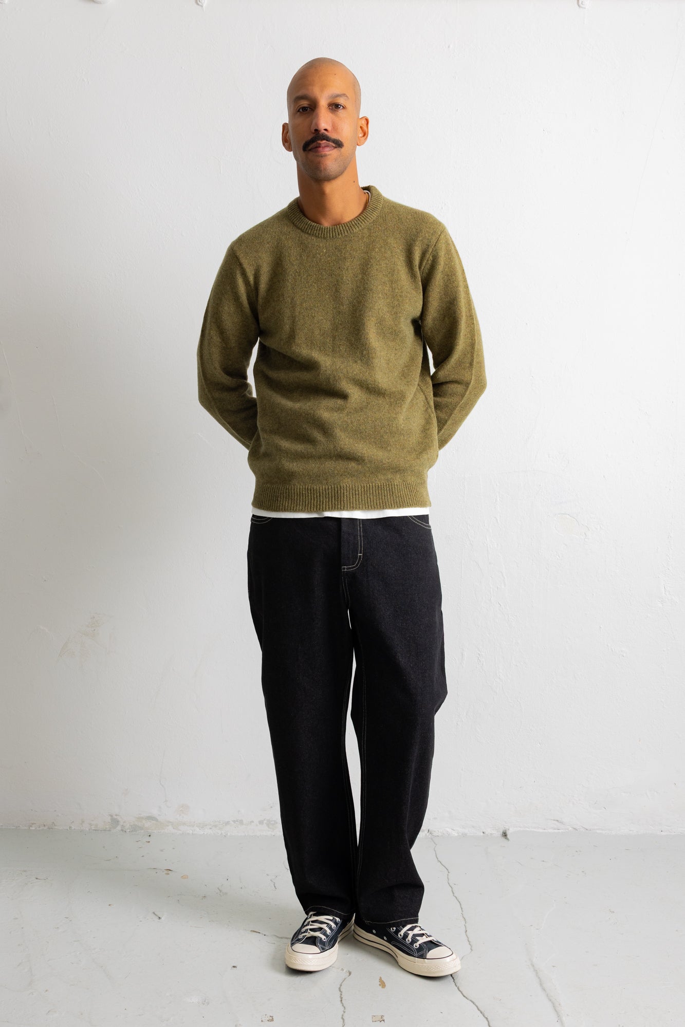 A man wearing an olive green crewneck sweater, black pants, and black-and-white sneakers, standing against a plain white wall.