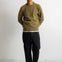 A man wearing an olive green crewneck sweater, black pants, and black-and-white sneakers, standing against a plain white wall.