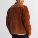 A man viewed from the back wearing a brown corduroy jacket and dark navy pants, standing against a plain light gray wall.