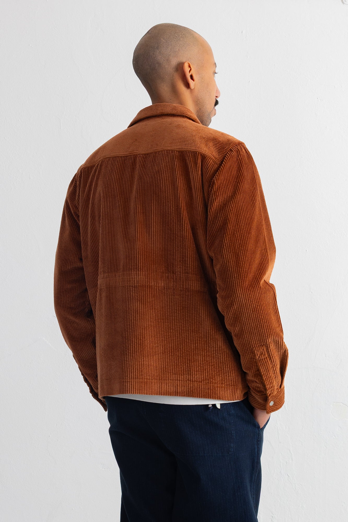 A man viewed from the back wearing a brown corduroy jacket and dark navy pants, standing against a plain light gray wall.