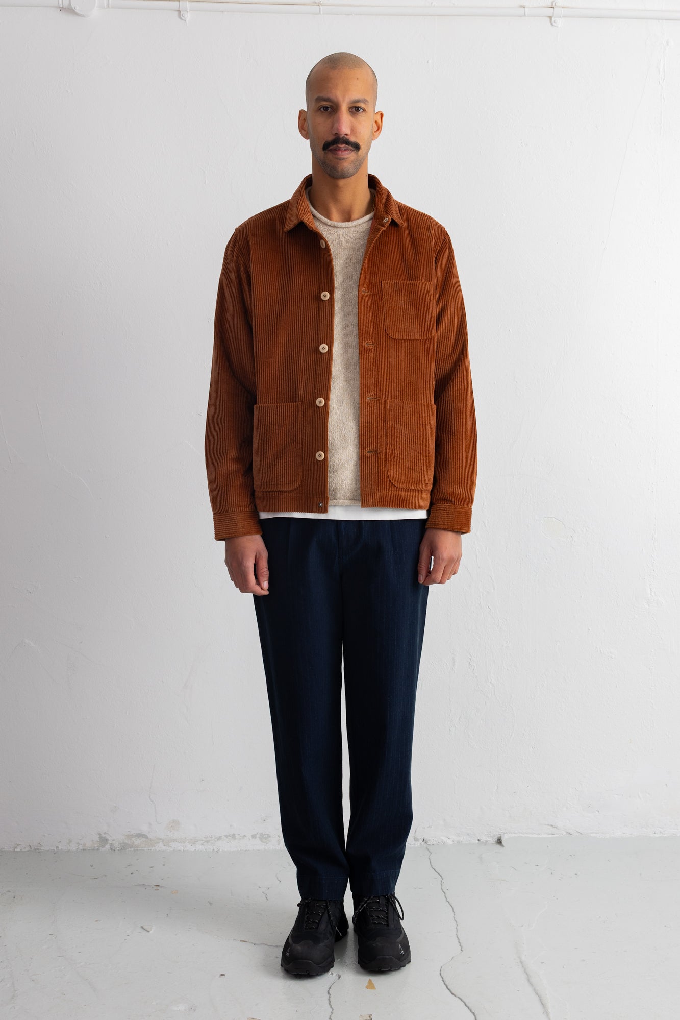 A man wearing a brown corduroy jacket over a cream shirt and navy pants, standing against a white wall.