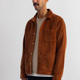 A man wearing a brown corduroy jacket over a beige t-shirt and dark blue pants, standing against a white wall.