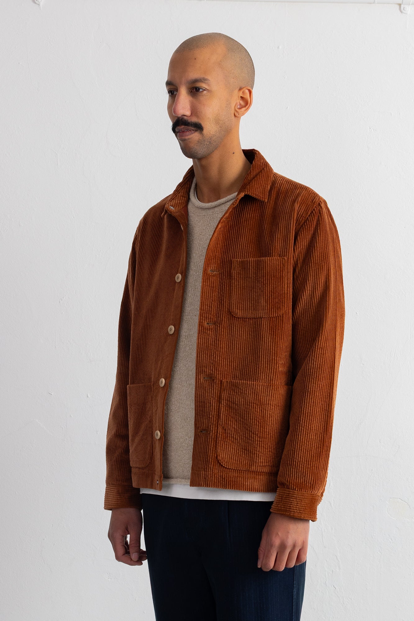 A man wearing a brown corduroy jacket over a beige t-shirt and dark blue pants, standing against a white wall.