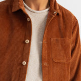 A close-up of a man wearing a rust-brown corduroy button-up shirt over a light beige knit sweater, against a plain off-white background.