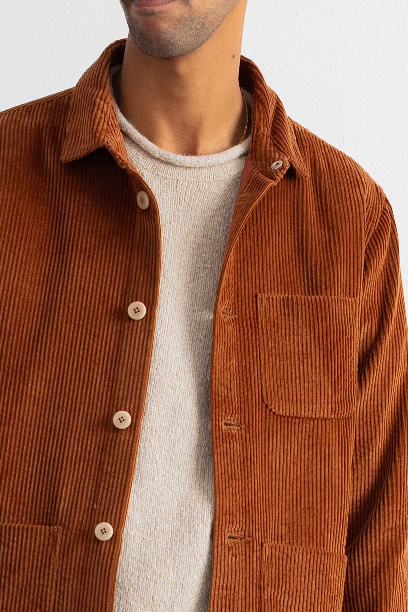 A close-up of a man wearing a rust-brown corduroy button-up shirt over a light beige knit sweater, against a plain off-white background.