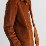 A man wearing a rust-brown corduroy button-up shirt jacket over a white t-shirt and navy blue trousers, shown from the chest down against a plain white wall.