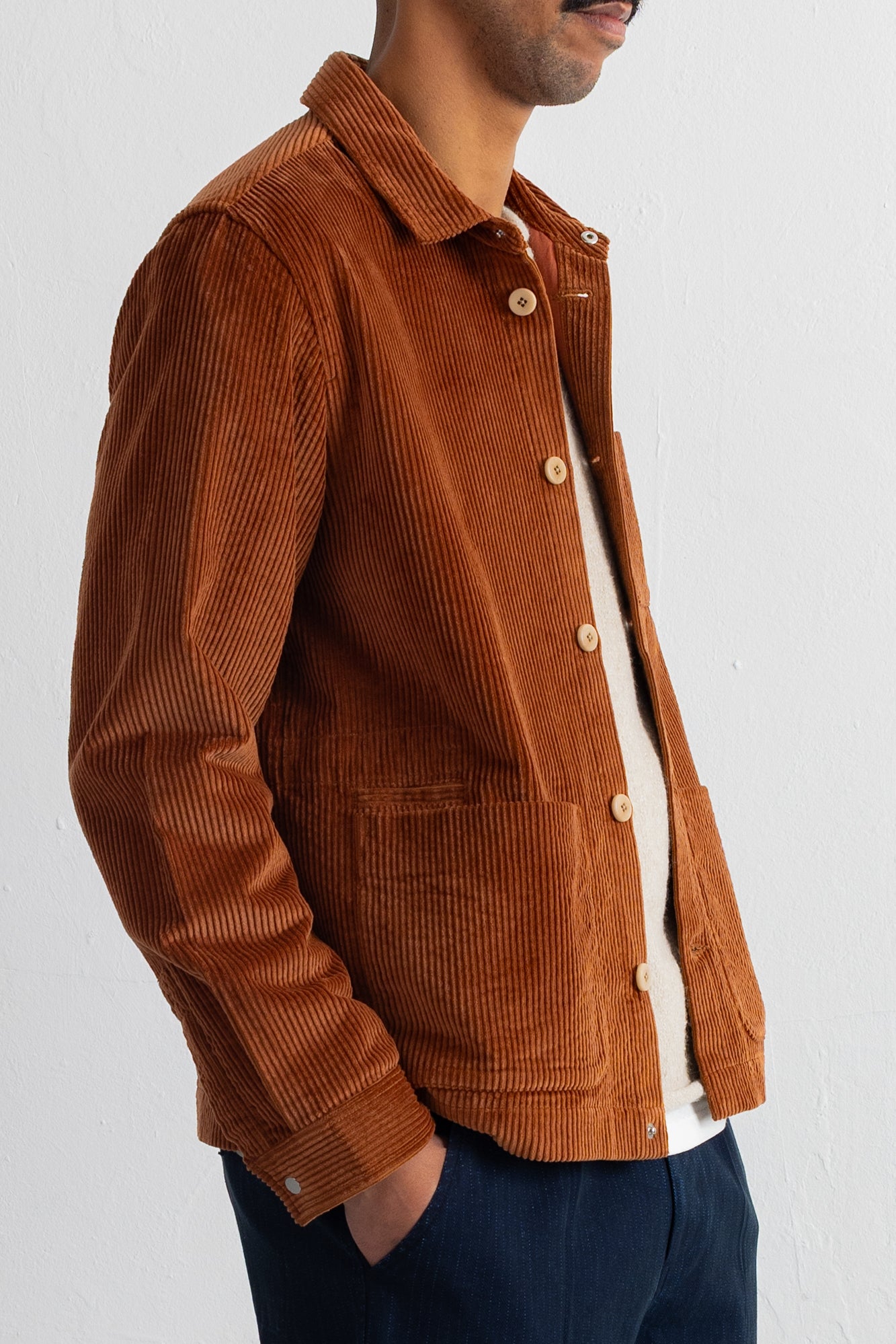A man wearing a rust-brown corduroy button-up shirt jacket over a white t-shirt and navy blue trousers, shown from the chest down against a plain white wall.