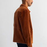 A man shown in profile wearing a brown corduroy jacket and dark pants, standing against a plain white wall.