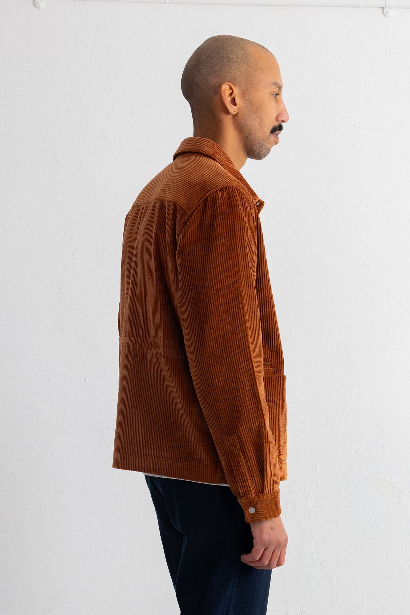 A man shown in profile wearing a brown corduroy jacket and dark pants, standing against a plain white wall.