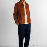 A man wearing a brown corduroy jacket over a white t-shirt, navy blue pants, and black shoes, standing against a white studio wall.