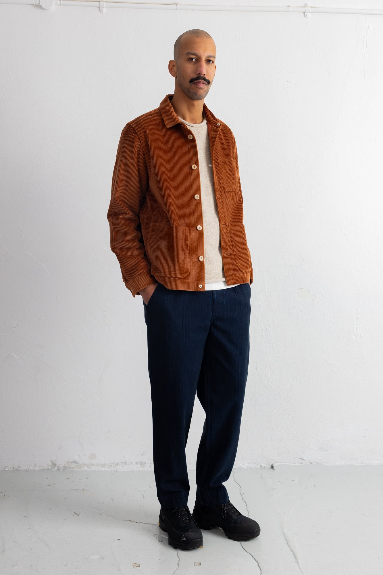 A man wearing a brown corduroy jacket over a white t-shirt, navy blue pants, and black shoes, standing against a white studio wall.