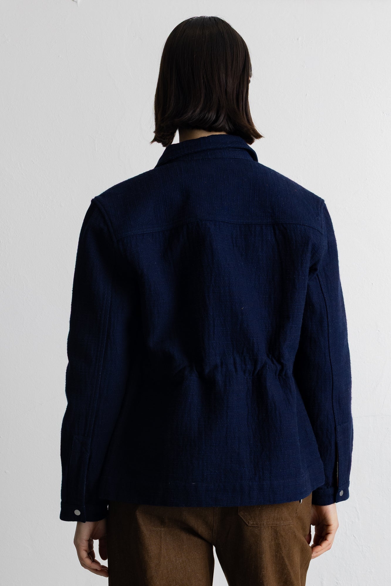 A person shown from the back wearing a dark navy blue textured jacket and brown pants against a plain white wall.