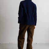 A person shown from the back wearing a dark navy blue jacket, brown pants, and beige boots, standing against a plain white wall.