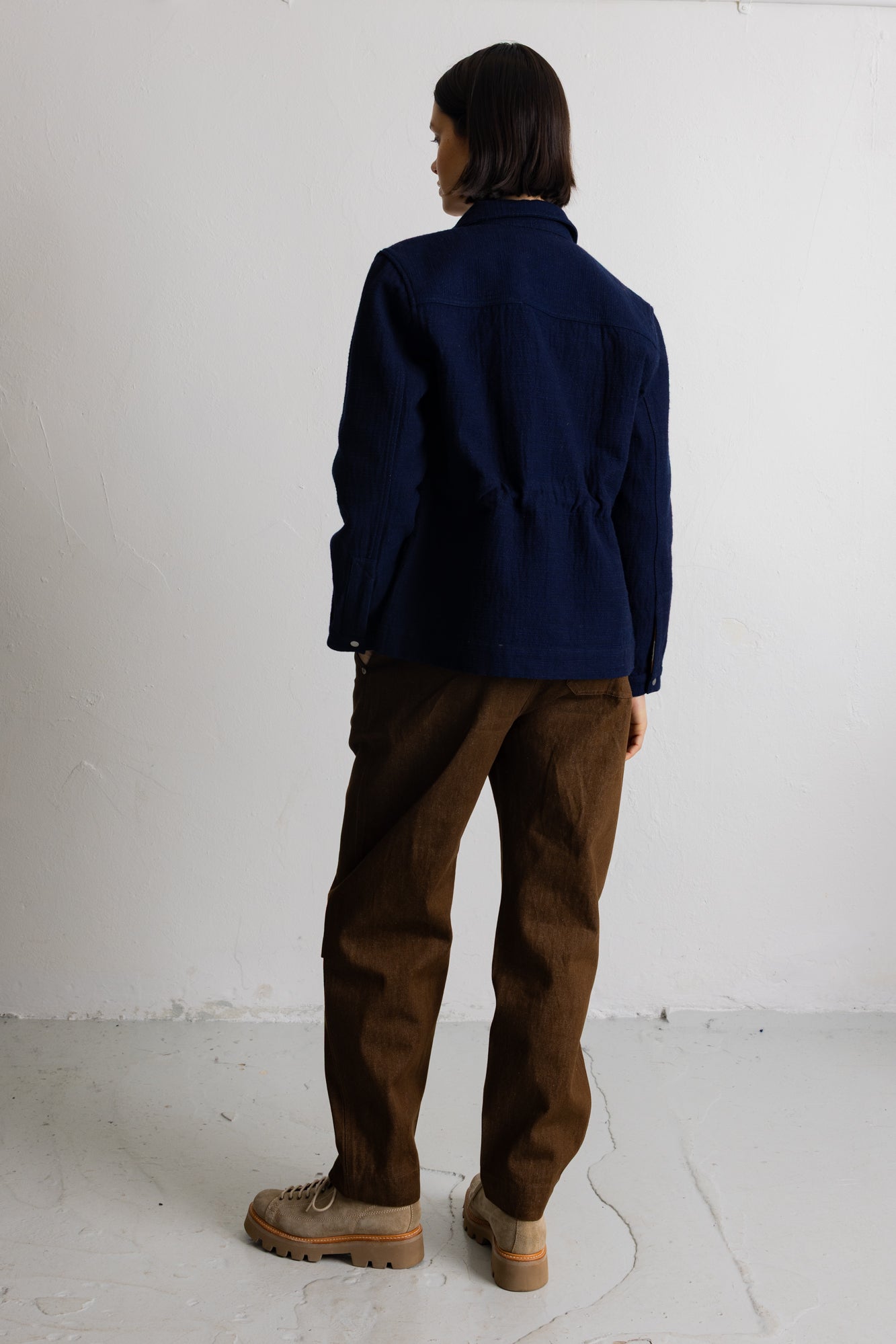 A person shown from the back wearing a dark navy blue jacket, brown pants, and beige boots, standing against a plain white wall.