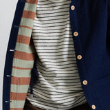 A close-up of a person wearing a navy blue button-front overshirt with a striped knit shirt underneath and brown trousers. The overshirt has visible silver metal snap buttons and a striped lining in muted green, peach, and navy.