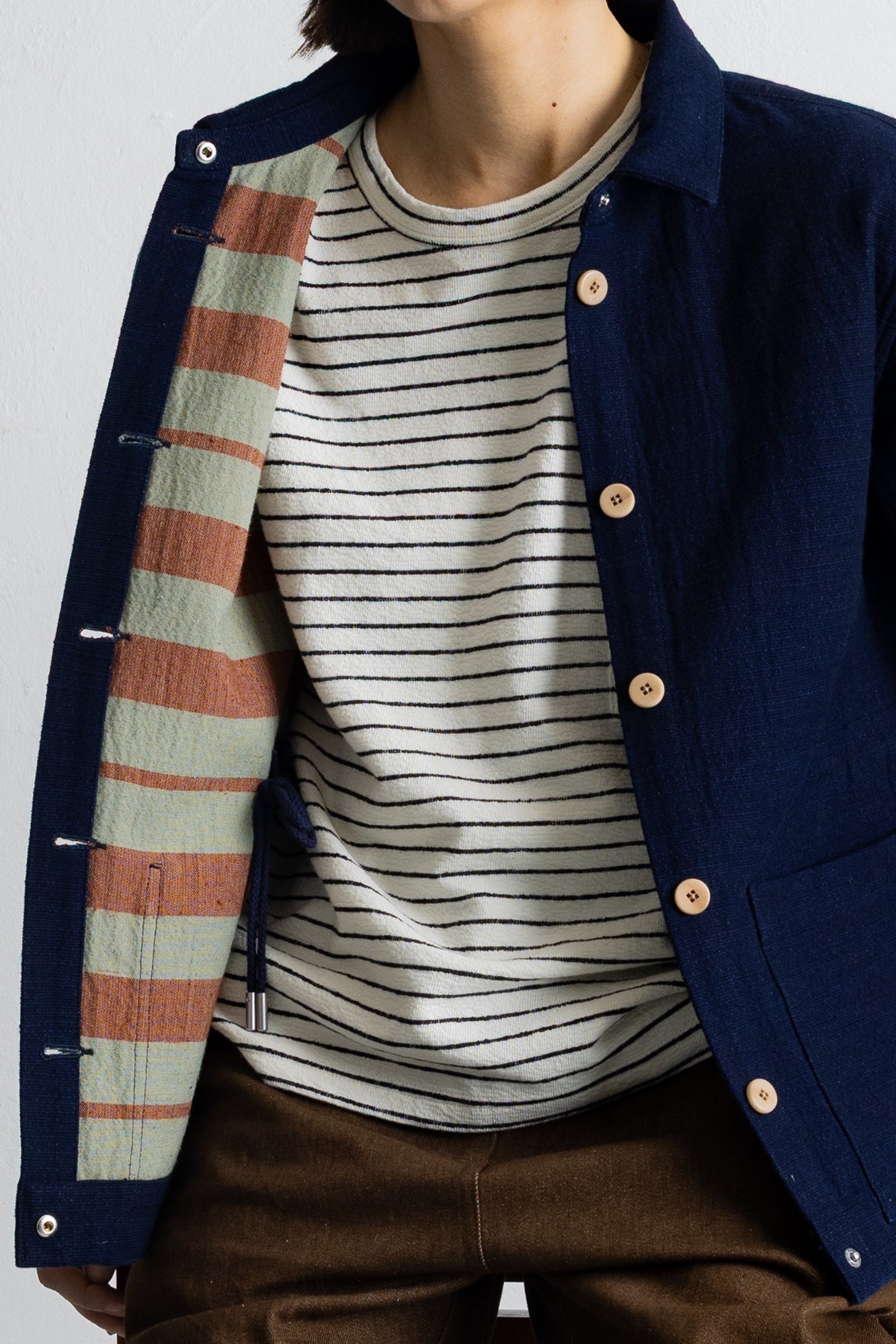 A close-up of a person wearing a navy blue button-front overshirt with a striped knit shirt underneath and brown trousers. The overshirt has visible silver metal snap buttons and a striped lining in muted green, peach, and navy.
