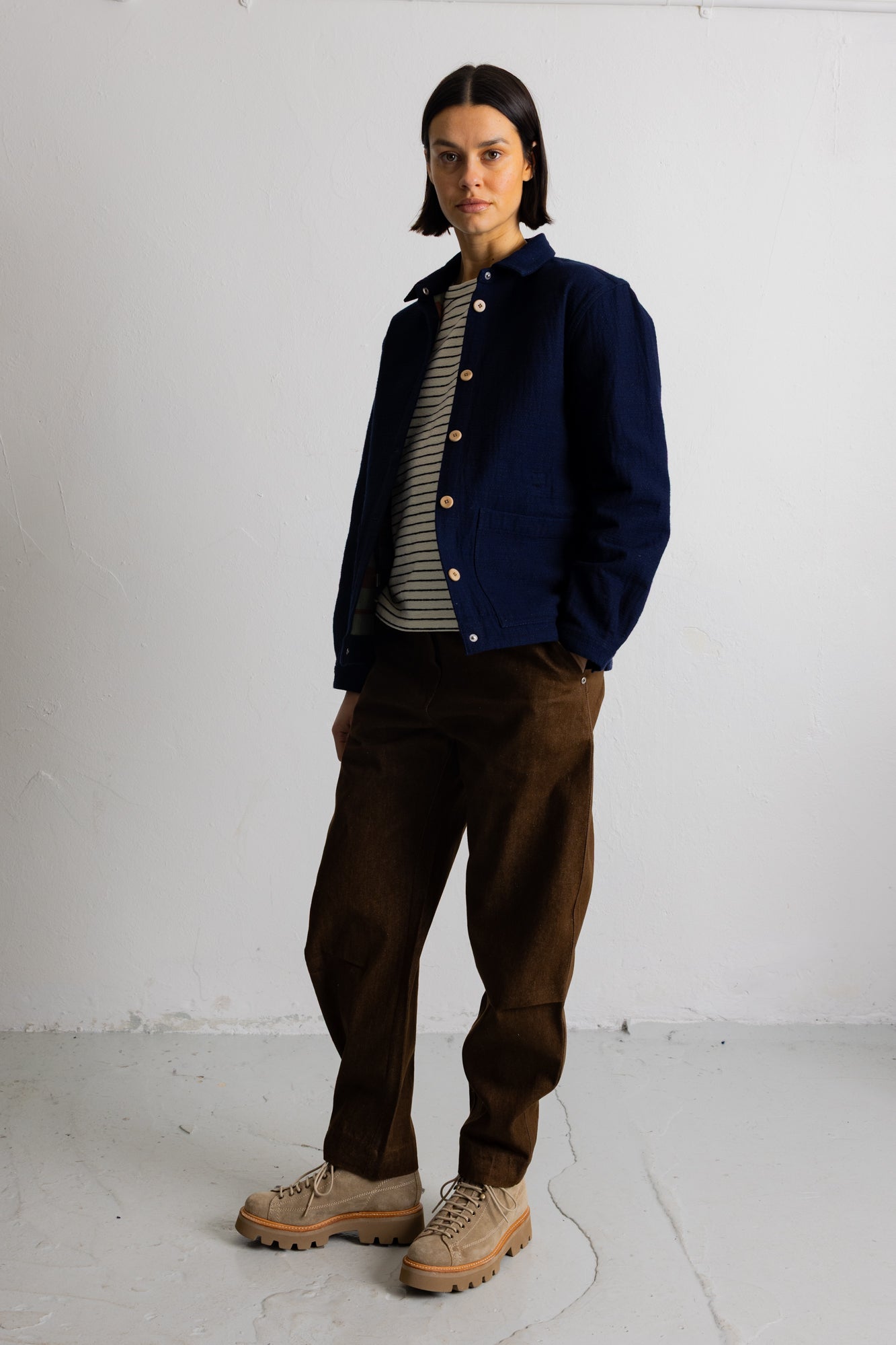A model stands against a white studio wall wearing a navy blue button-up jacket over a gray and white striped shirt, paired with brown corduroy pants and beige lace-up boots.