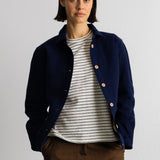 A model wearing a navy blue corduroy jacket over a white and black striped t-shirt, paired with brown trousers, standing against a plain light gray wall.