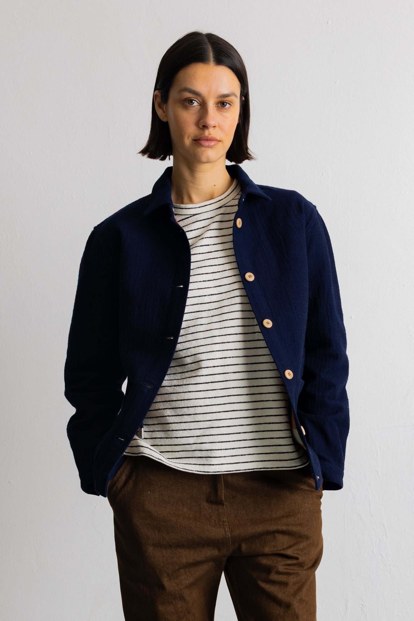 A model wearing a navy blue corduroy jacket over a white and black striped t-shirt, paired with brown trousers, standing against a plain light gray wall.