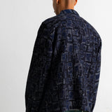 A man viewed from behind wearing a dark navy blue patterned button-up shirt and teal-green jeans against a plain white background.