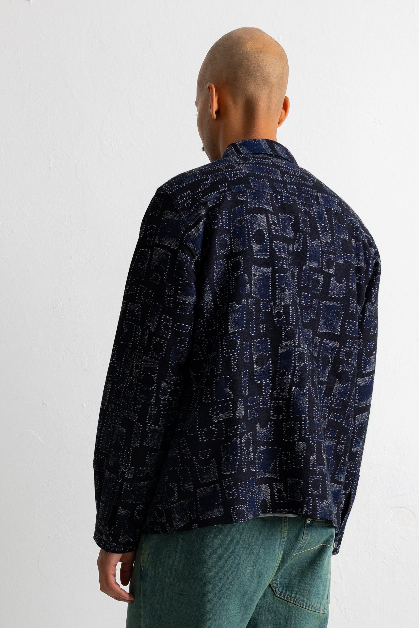 A man viewed from behind wearing a dark navy blue patterned button-up shirt and teal-green jeans against a plain white background.