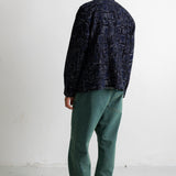 A man is shown from behind wearing a dark navy blue patterned shirt, green pants, and black canvas sneakers, standing against a plain white wall.