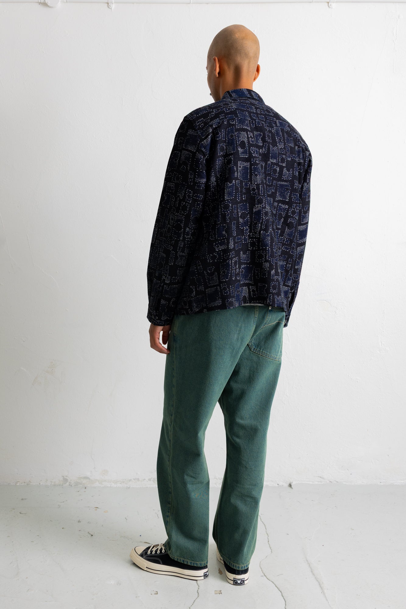 A man is shown from behind wearing a dark navy blue patterned shirt, green pants, and black canvas sneakers, standing against a plain white wall.