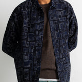 A man wearing a dark navy blue patterned button-up shirt jacket over a brown knit sweater and teal pants, shown from the chin down against a plain white wall.