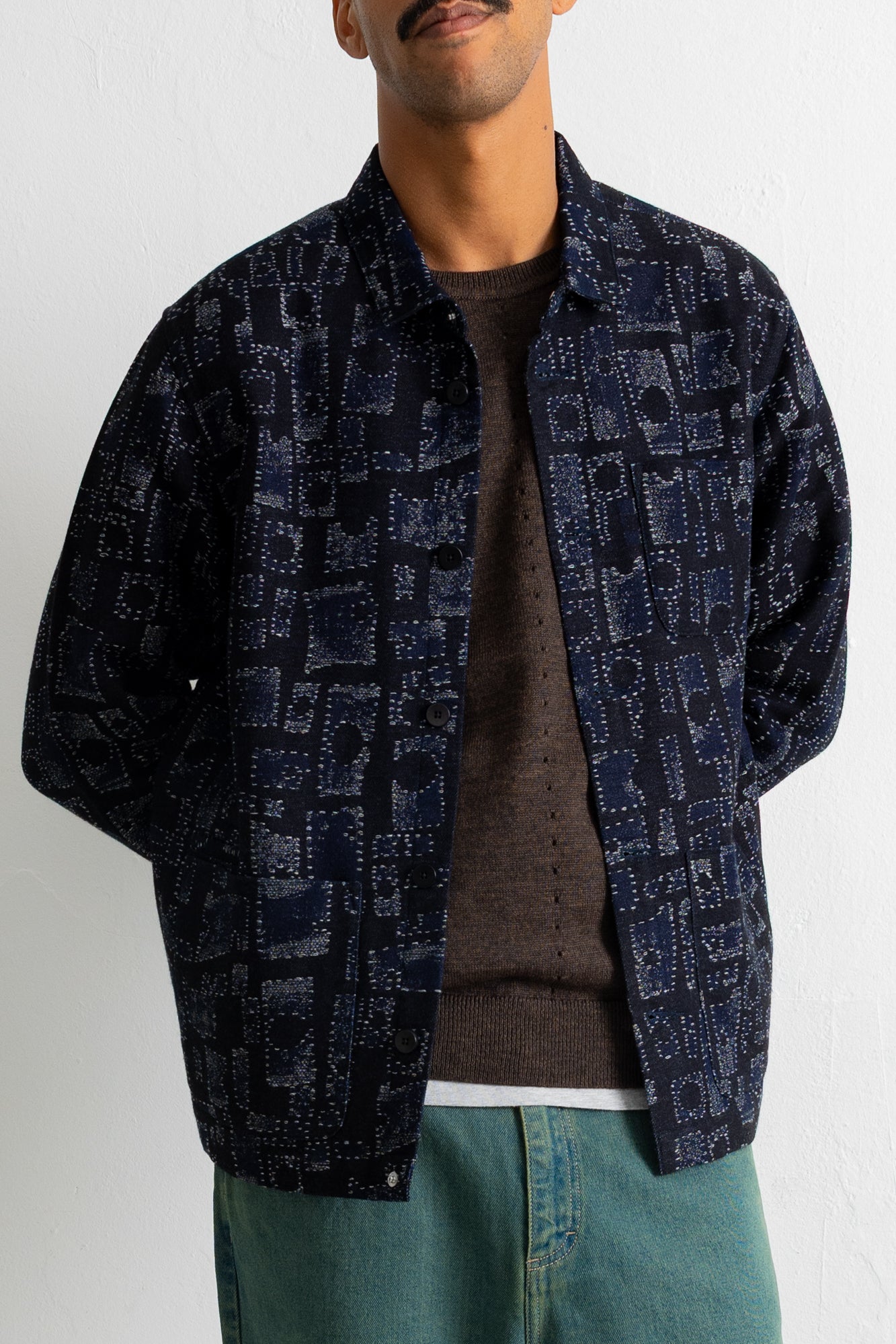 A man wearing a dark navy blue patterned button-up shirt jacket over a brown knit sweater and teal pants, shown from the chin down against a plain white wall.