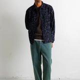 A man stands against a white wall wearing a dark navy patterned overshirt over a dark brown t-shirt, green pants, and black and white sneakers.